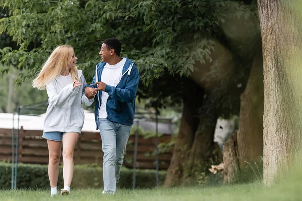 Comprimento total de feliz casal multiétnico andando e falando no parque — Fotografia de Stock