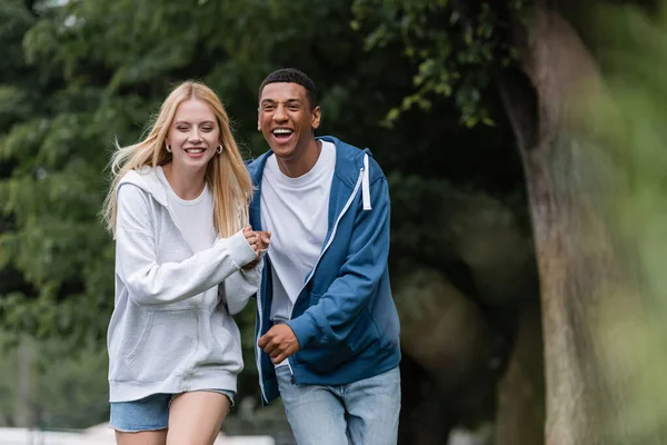 Femme blonde heureuse et rire homme afro-américain tenant la main et marchant dans le parc — Photo de stock