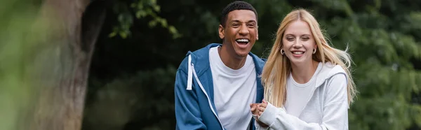 Alegre africano americano hombre riendo cerca joven rubia novia en parque, bandera - foto de stock