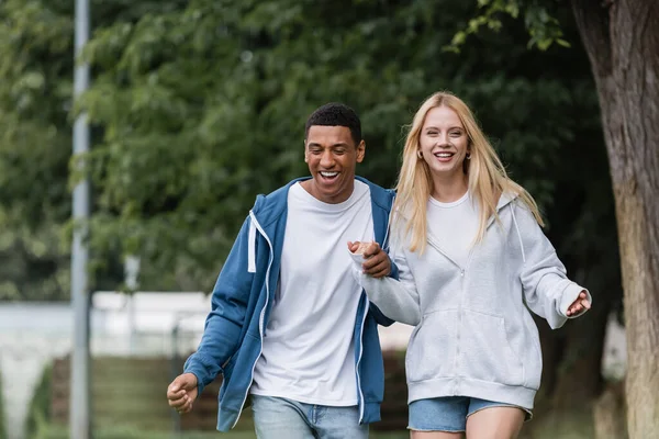 Cheerful multiethnic couple in hoodies holding hands while walking in green park — Stock Photo
