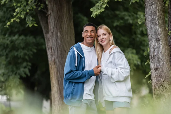 Complacida pareja interracial cogida de la mano y mirando hacia otro lado mientras está de pie en el parque - foto de stock