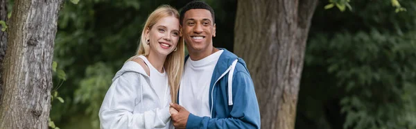 Casal interracial positivo em capuzes sorrindo e de mãos dadas no parque, banner — Fotografia de Stock