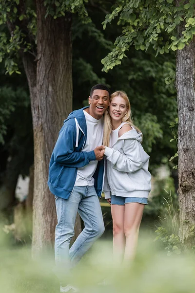 Gioioso afroamericano uomo e giovane donna bionda con cappuccio tenendosi per mano e guardando la fotocamera nel parco — Foto stock
