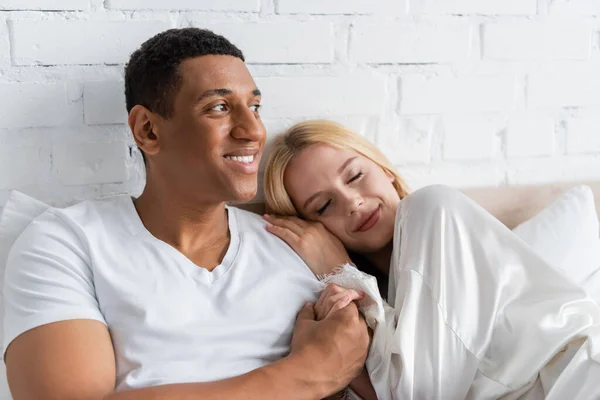 Mujer sonriente con los ojos cerrados apoyados en el hombro del hombre afroamericano feliz sosteniendo su mano y mirando hacia otro lado - foto de stock