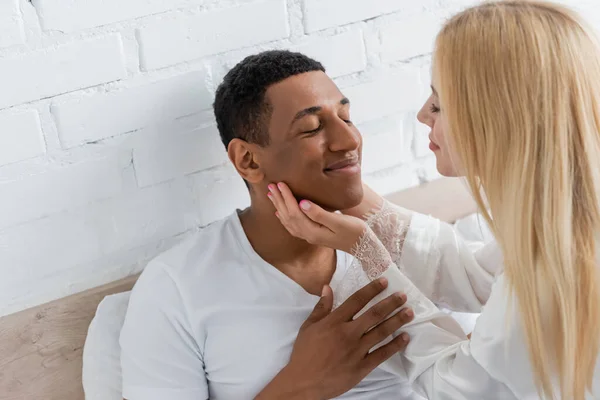Jeune femme blonde touchant le visage du petit ami afro-américain souriant avec les yeux fermés dans la chambre — Photo de stock