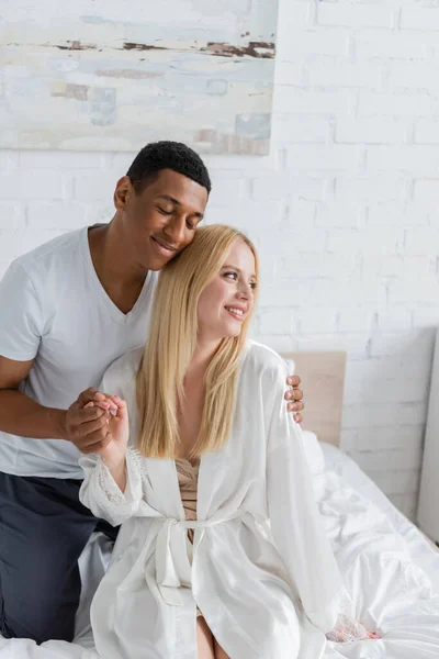 Tierno afroamericano hombre sonriendo con los ojos cerrados mientras toma de la mano de su novia rubia y abrazándola en el dormitorio - foto de stock