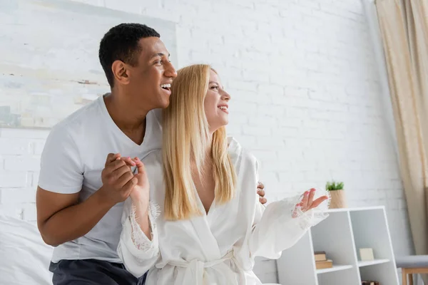 Hombre afroamericano feliz y mujer rubia en túnica blanca tomados de la mano y mirando hacia otro lado en el dormitorio - foto de stock