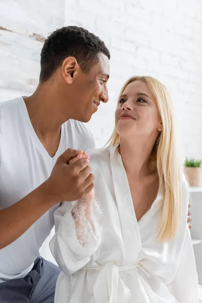 Feliz pareja multiétnica en ropa de dormir blanca tomados de la mano y mirándose en el dormitorio - foto de stock