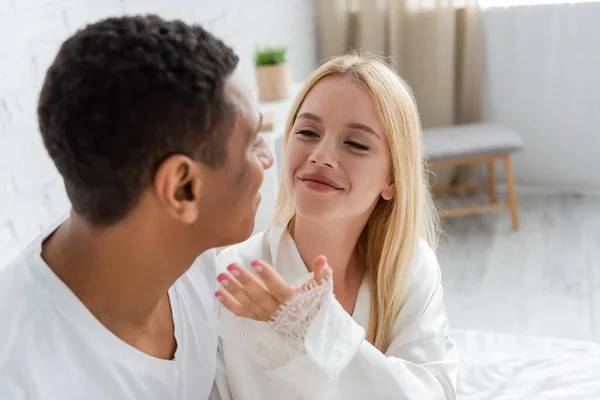 Happy young woman holding hand near blurred african american man in bedroom — Stock Photo