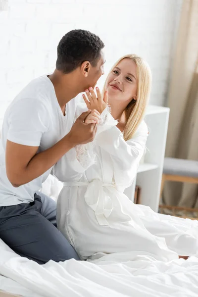 Femme souriante en robe blanche touchant le visage du petit ami afro-américain tenant sa main dans la chambre — Photo de stock