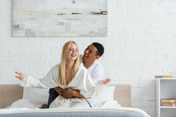 Glücklicher afrikanisch-amerikanischer Mann umarmt Taille einer Frau im weißen Gewand, die mit geschlossenen Augen auf dem Bett sitzt — Stockfoto