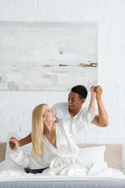 Joyeux couple interracial tenant la main et souriant à l'autre tout en s'amusant dans la chambre — Photo de stock