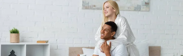 Young blonde woman in white robe embracing african american man and looking away in bedroom, banner — Stock Photo