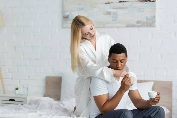 African american man sitting on bed with coffee cup and kissing hand of smiling girlfriend in white silk robe — Stock Photo