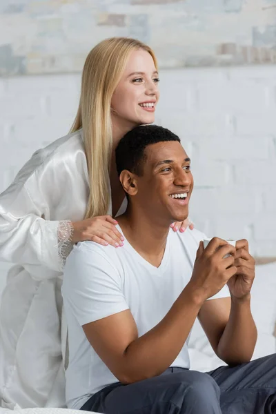 Mujer rubia en túnica de seda blanca y alegre hombre afroamericano con taza de café mirando hacia otro lado en el dormitorio - foto de stock