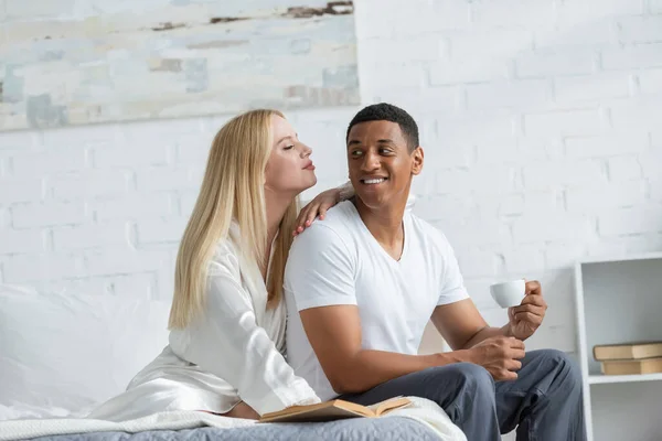 Happy african american man looking at blonde girlfriend in white silk robe while sitting on bed with coffee cup — Stock Photo