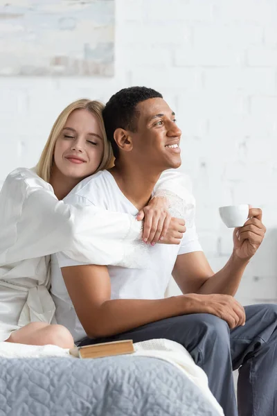 Jovem mulher loira com olhos fechados abraçando feliz namorado afro-americano segurando xícara de café no quarto — Fotografia de Stock