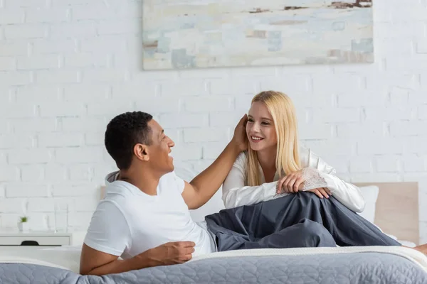 Glücklich afrikanisch-amerikanischer Mann berührt Haare der jungen blonden Freundin auf dem Bett in der Früh — Stockfoto
