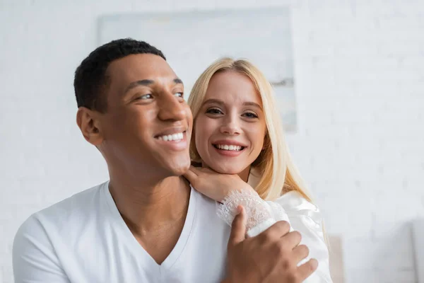 Femme blonde gaie appuyée sur l'épaule du petit ami afro-américain et regardant la caméra dans la chambre — Photo de stock