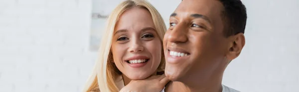 Jovem e alegre mulher olhando para a câmera perto de homem afro-americano sorrindo em primeiro plano borrado, bandeira — Fotografia de Stock