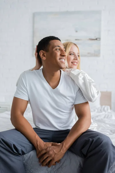 Homem americano africano em t-shirt branca e calças de pijama sentado na cama perto de namorada loira e olhando para longe — Fotografia de Stock