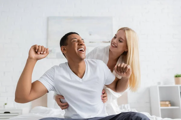Cheerful woman tickling african american boyfriend laughing in bedroom — Stock Photo