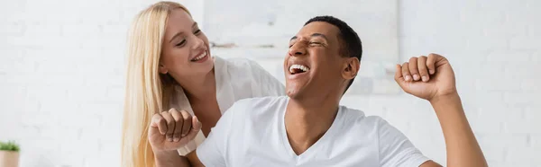 Hombre afroamericano emocionado con los ojos cerrados riendo cerca de la mujer rubia sonriente en el dormitorio, pancarta - foto de stock