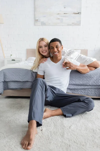 Descalzo afroamericano hombre en pantalones de pijama sentado en el suelo cerca de novia en la cama y mirando a la cámara - foto de stock