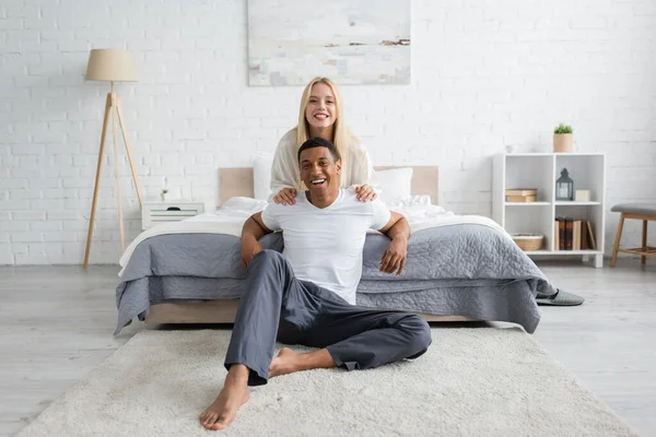 Cheerful african american man in sleepwear sitting on floor in bedroom near blonde woman hugging his shoulders — Stock Photo