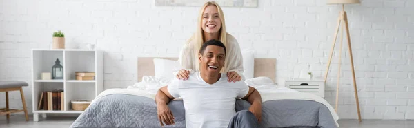 Alegre pareja multiétnica en ropa de casa sonriendo a la cámara en amplio dormitorio, pancarta - foto de stock