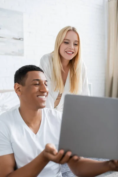 Happy african american man showing laptop to young smiling girlfriend in bedroom — Stock Photo