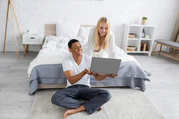 African american man sitting on floor and holding laptop near blonde girlfriend in bedroom — Stock Photo