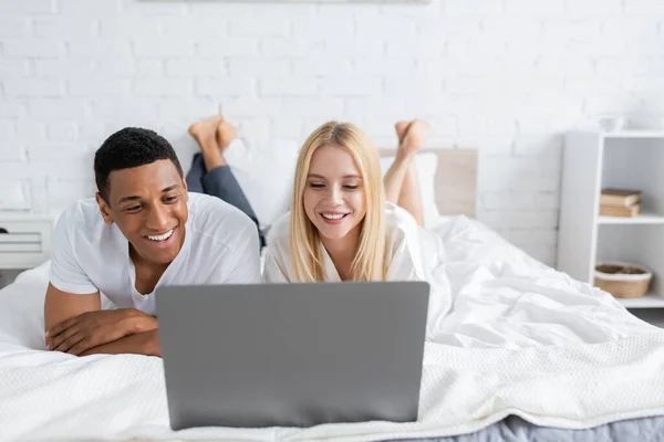 Donna bionda con fidanzato afroamericano che guarda film comico sul computer portatile sul letto — Foto stock