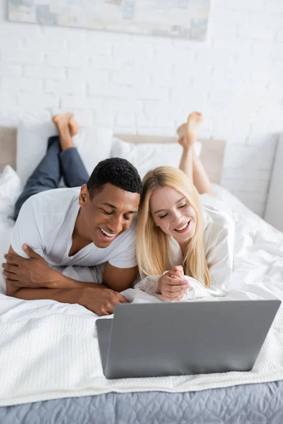 Босоногая межрасовая пара улыбается во время просмотра фильма на ноутбуке в спальне — стоковое фото