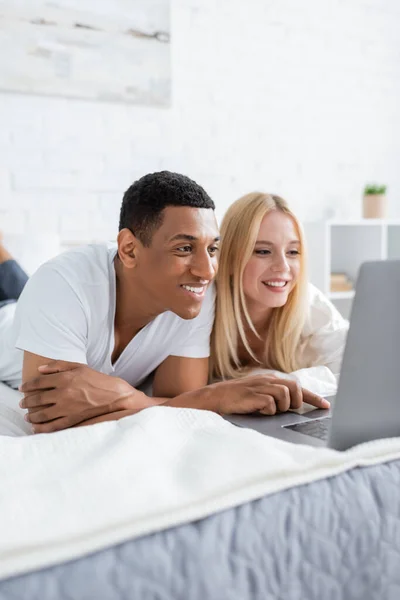 Щаслива багатоетнічна пара лежить на ліжку і дивиться фільм на ноутбуці — стокове фото