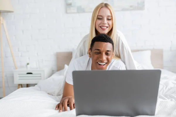 Веселий афроамериканський чоловік дивиться комедію на ноутбук біля усміхненої дівчини в спальні — стокове фото