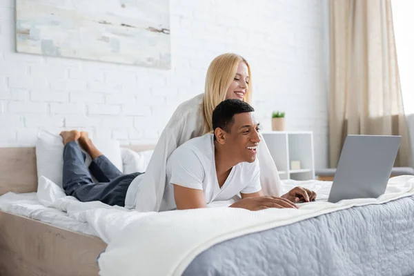 Молодая блондинка улыбается рядом с веселым африканским американским парнем, смотрящим фильм на ноутбуке в спальне — стоковое фото