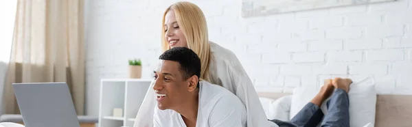 Junge blonde Frau und fröhliche afrikanisch-amerikanische Mann beobachten Komödie auf Laptop im Schlafzimmer, Banner — Stockfoto