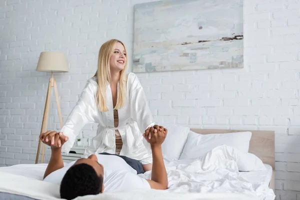 Mujer feliz en túnica de seda blanca mirando hacia otro lado y tomándose de la mano con el hombre afroamericano acostado en la cama - foto de stock