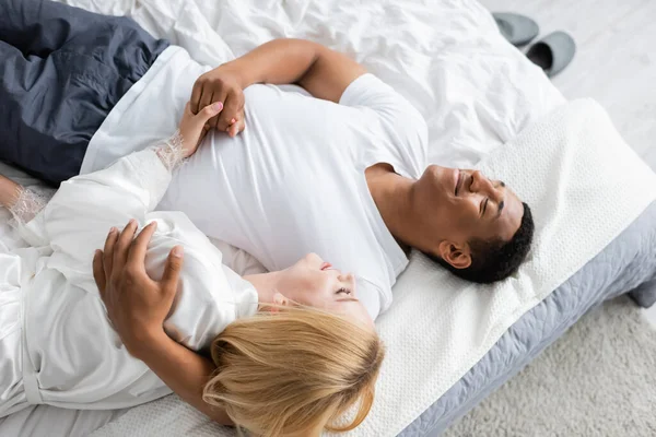 Top view of african american man with closed eyes hugging blonde woman and holding her hand on bed — Stock Photo