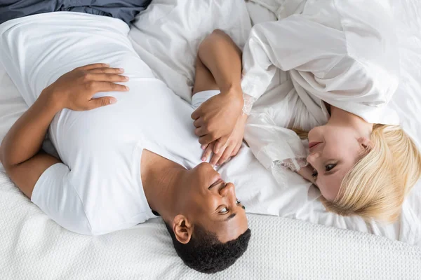 Vista superior do casal multiétnico satisfeito de mãos dadas e olhando um para o outro na cama — Fotografia de Stock