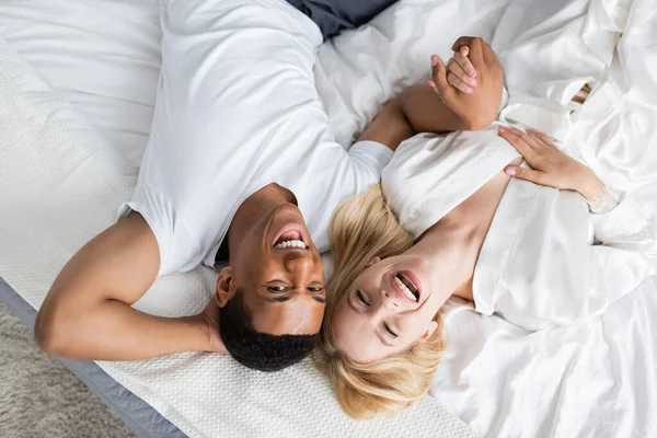 Vista superior de pareja multiétnica emocionada tomados de la mano y riendo de la cámara en la ropa de cama blanca - foto de stock