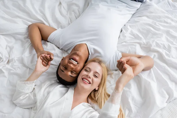 Vista superior de hombre afroamericano feliz en camiseta y mujer en bata de seda acostado con los ojos cerrados y cogido de la mano - foto de stock