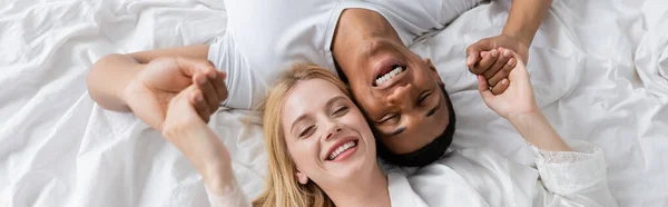 Vista superior de alegre pareja interracial en el amor acostado sobre ropa de cama blanca con los ojos cerrados y cogidas de la mano, pancarta - foto de stock