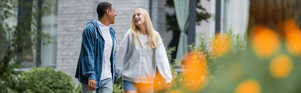 Happy blonde woman talking to african american boyfriend while walking in city on blurred foreground, banner — Stock Photo