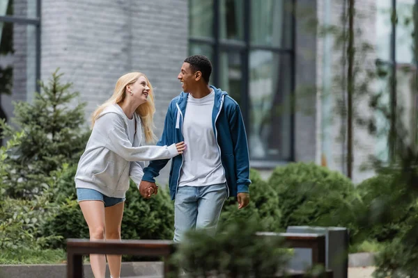 Радісна багатоетнічна пара тримає руки і посміхається один одному під час прогулянки по міській вулиці — стокове фото