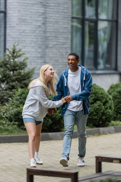 Full length of cheerful interracial couple walking and laughing on urban street — Stock Photo