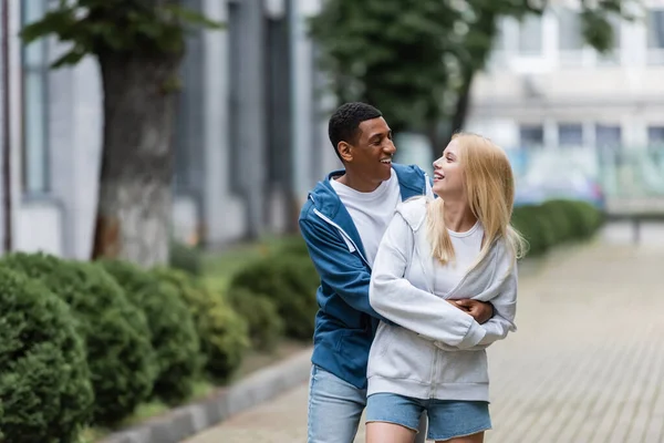 Gioioso africano americano uomo abbracciando giovane bionda fidanzata su strada sfocata in città — Foto stock