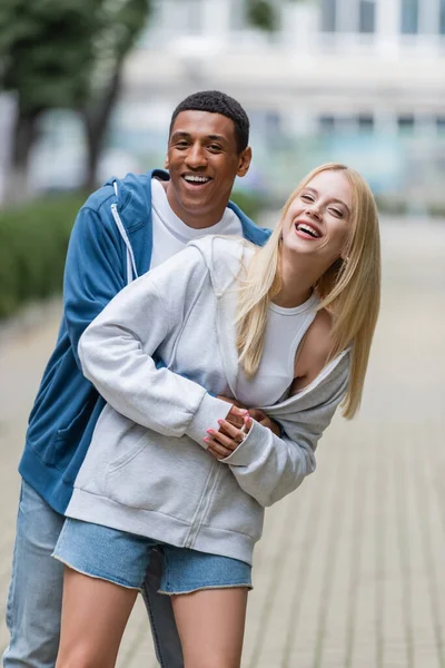 Mujer rubia feliz con novio afroamericano en sudaderas con capucha mirando a la cámara en la calle borrosa - foto de stock