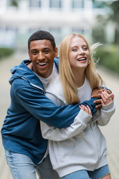 African american man laughing and hugging blonde girlfriend on blurred street in city — Stock Photo